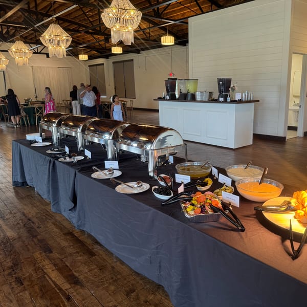 A buffet line with chafing dishes, salads, dressings, and a drink station in a large event space.